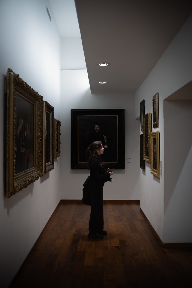 Visiteuse de profil dans une salle de musée, entre des tableaux aux cadres dorés, sous un puits de lumière