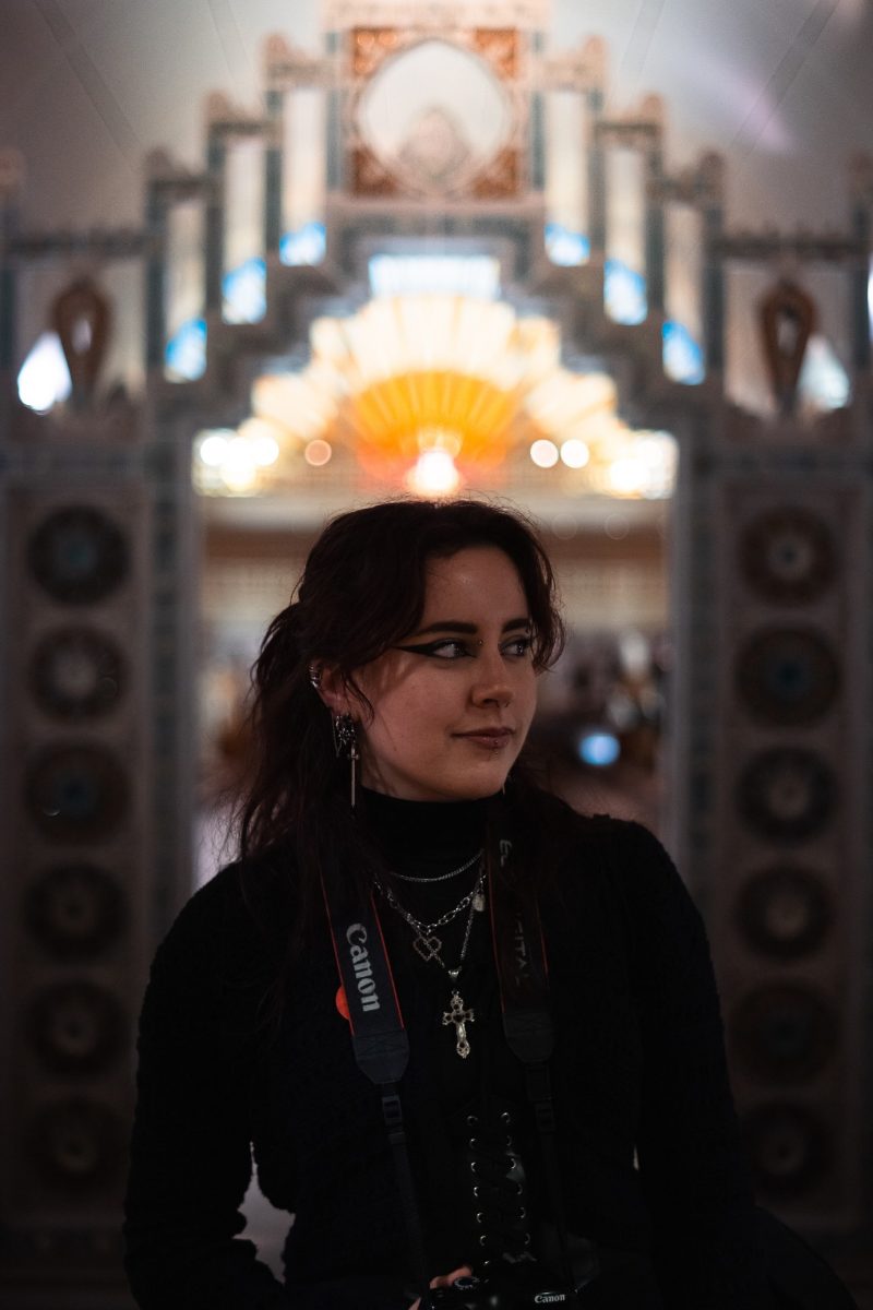 Portrait d'une jeune femme aux cheveux bruns, appareil photo Canon en bandoulière, devant un décor Art déco lumineux et flou