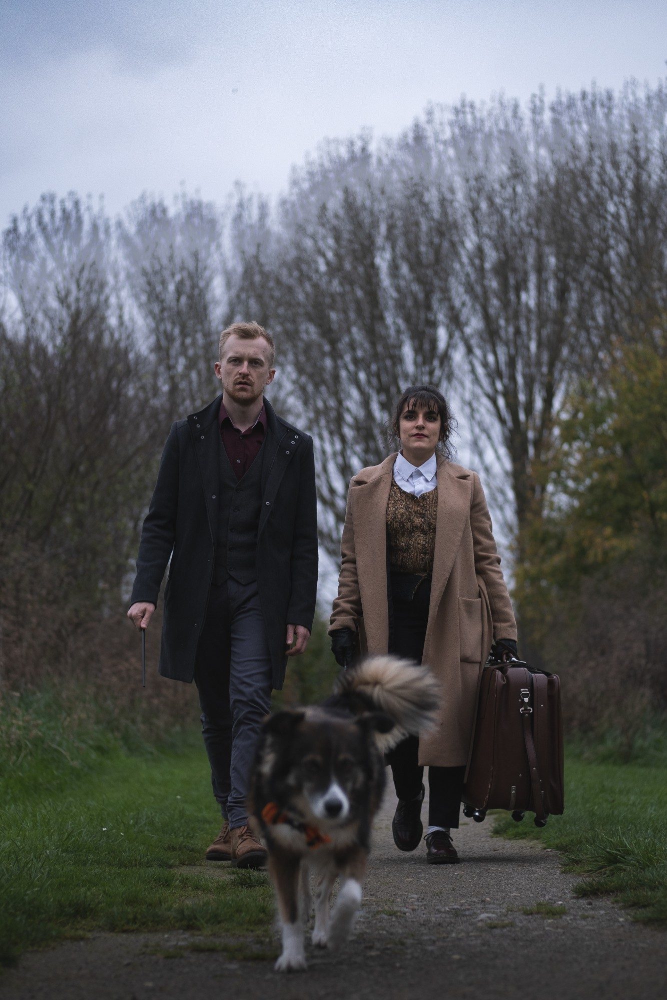 Couple marchant vers l'objectif sur un chemin de campagne, baguette et valise ancienne à la main, précédé d'un chien à poil long